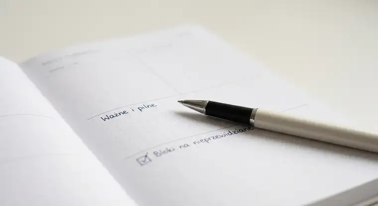Pracownik w korporacyjnym open space planuje dzień w papierowym plannerze dziennym, próbując uporządkować zadania i priorytety w hałaśliwym biurze